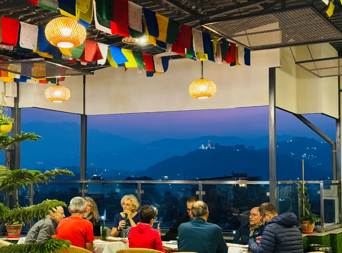 Diners under a string of prayer flags on the rooftop at dusk, the Kathmandu hills and the lit Swayambhunath stupa beyond