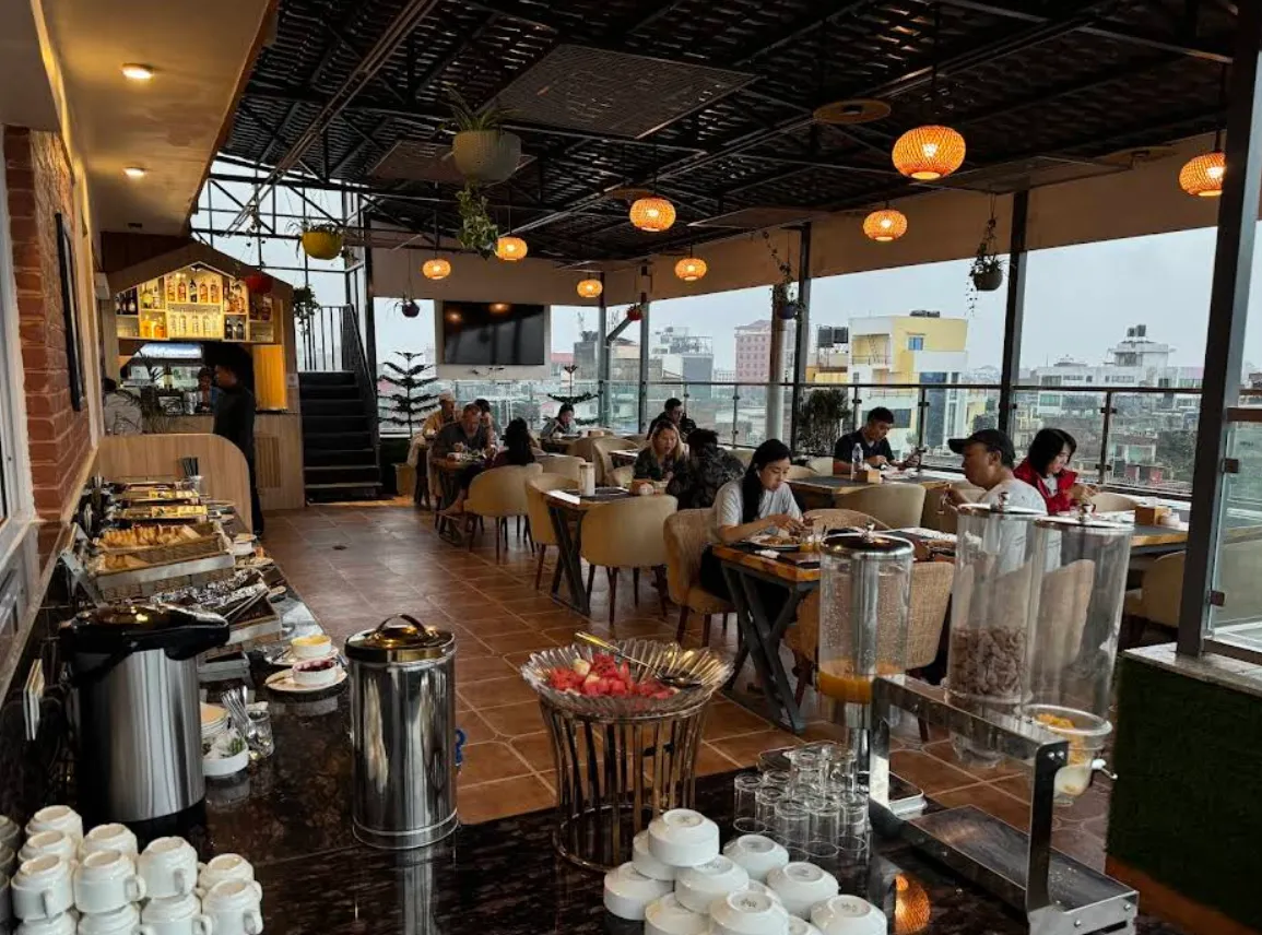 The two-level rooftop interior at golden hour — open kitchen and buffet, warm lanterns, diners and the city through the glass