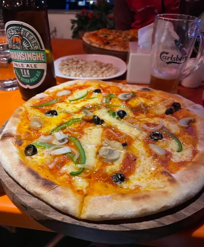 A wood-fired pizza on a board beside cold bottles at night